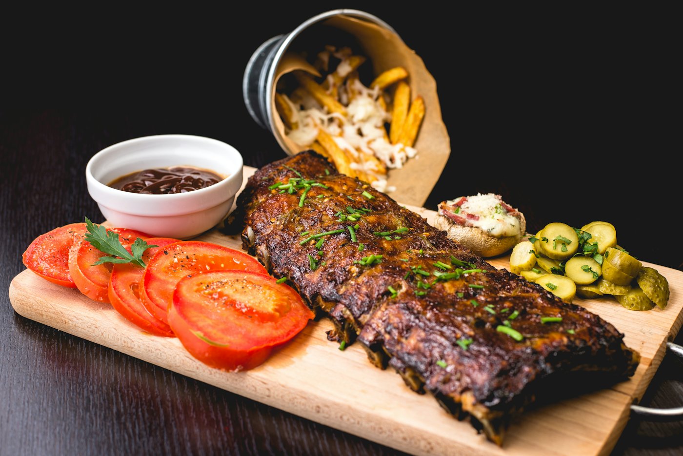 Travers de porc laqués sur une planche, tomates et frites, sur fond noir